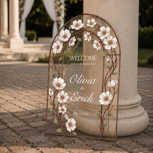 K&ouml;nnte beinhalten: Ein bogenf&ouml;rmiges Hochzeits-Willkommensschild mit einem braunen Metallrahmen, verziert mit wei&szlig;en Blumenillustrationen und gr&uuml;nen Bl&auml;ttern. Der Text lautet: WELCOME we're so glad you're here, Olivia & Erick, sowie das Datum July 14, 2026.