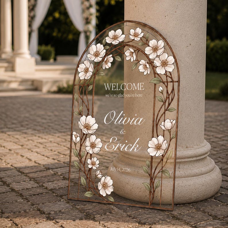 K&ouml;nnte beinhalten: Ein bogenf&ouml;rmiges Hochzeits-Willkommensschild mit einem braunen Metallrahmen, verziert mit wei&szlig;en Blumenillustrationen und gr&uuml;nen Bl&auml;ttern. Der Text lautet: WELCOME we're so glad you're here, Olivia & Erick, sowie das Datum July 14, 2026.