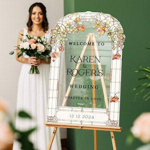 May include: An arched clear acrylic wedding sign featuring a printed stained glass border with autumn-colored leaves. The text reads: WELCOME TO KAREN & ROGER'S WEDDING FOREVER IN LOVE 12 12 2024. The sign is displayed on a light wood tripod easel.