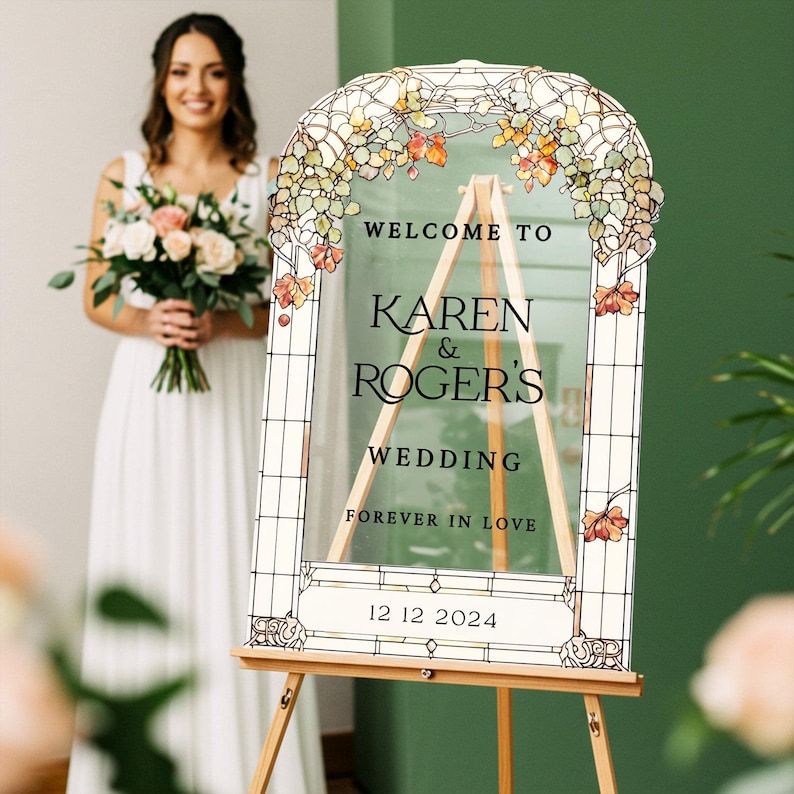 May include: An arched clear acrylic wedding sign featuring a printed stained glass border with autumn-colored leaves. The text reads: WELCOME TO KAREN & ROGER'S WEDDING FOREVER IN LOVE 12 12 2024. The sign is displayed on a light wood tripod easel.