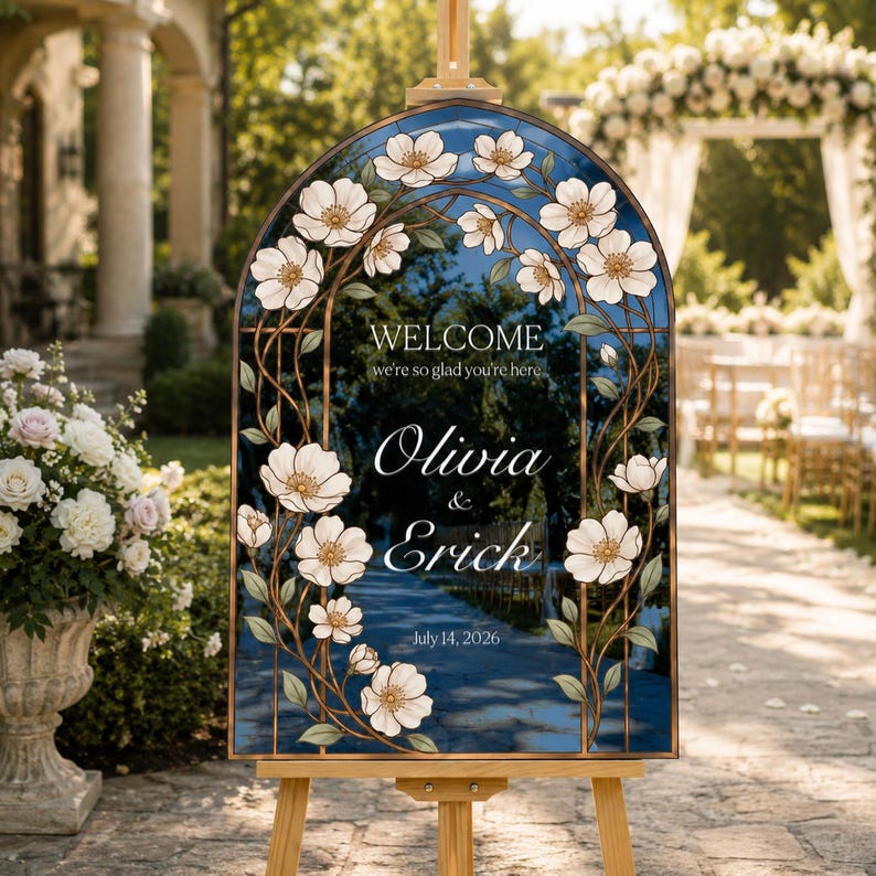 K&ouml;nnte beinhalten: Ein bogenf&ouml;rmiges Hochzeits-Willkommensschild mit dunkelblauem Hintergrund und einem wei&szlig;en Blumenrand aus illustrierten Bl&uuml;ten und Ranken. Der Text lautet: WELCOME we're so glad you're here, Olivia & Erick, July 14, 2026. Auf einem Holzst&auml;nder.
