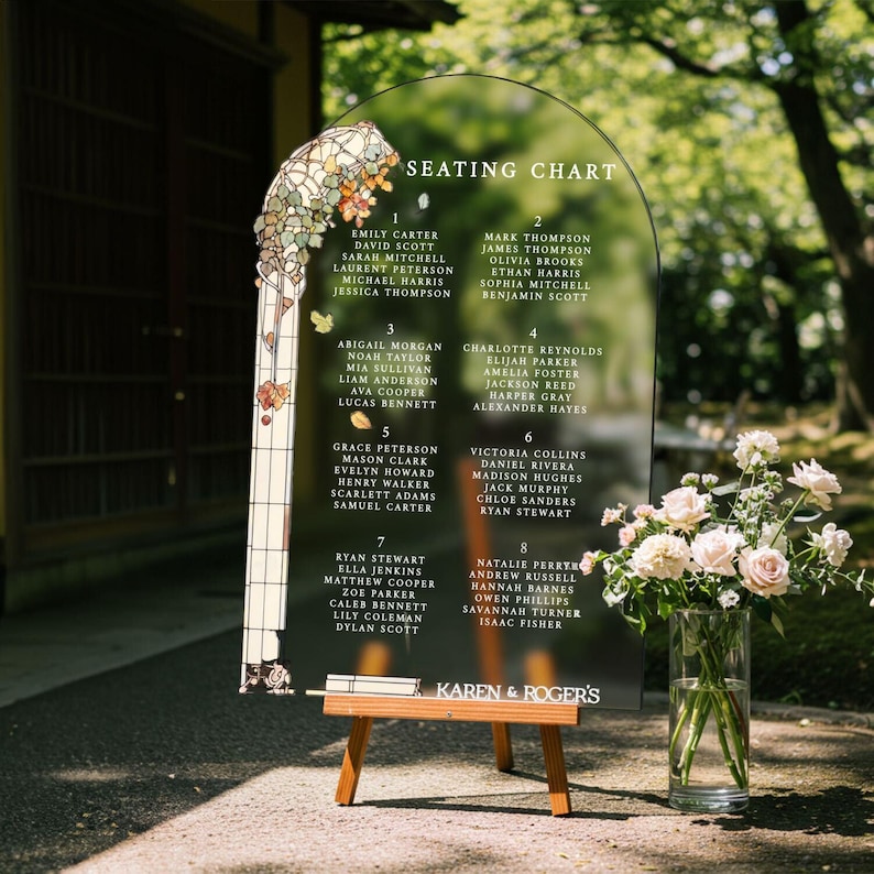 May include: An arched, transparent acrylic seating chart sign with white text listing names under eight numbered tables. The left side features a stained-glass style floral illustration. The sign rests on a wooden easel with the text KAREN & ROGER'S.