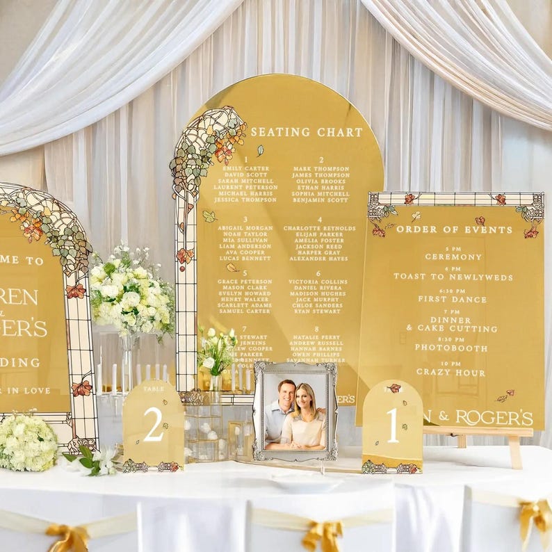 May include: A wedding seating chart and order of events sign with a gold mirror finish. The signs are decorated with floral and leaf designs. The seating chart lists guest names and table numbers. The order of events sign lists the ceremony, toast to newlyweds, first dance, dinner and cake cutting, photo booth, and crazy hour.