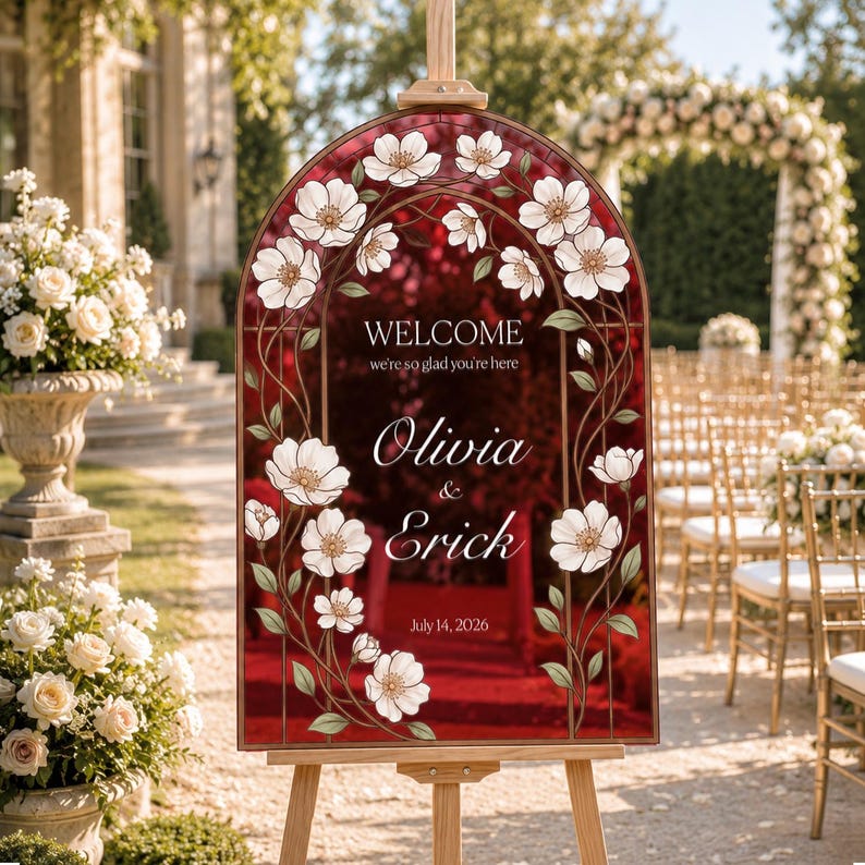 K&ouml;nnte beinhalten: Ein bogenf&ouml;rmiges Hochzeits-Willkommensschild mit tiefrotem Hintergrund und wei&szlig;em Blumenrand. Der Text lautet: WELCOME we're so glad you're here, Olivia & Erick, July 14, 2026. Das Schild steht auf einer h&ouml;lzernen Staffelei im Freien.