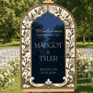 May include: An arched wedding welcome sign with a dark blue center panel featuring white text: Welcome THE WEDDING OF MARGOT & TYLER BOSTON, USA 07.15.2026. The frame is decorated with stained glass-style white flowers and green leaves.