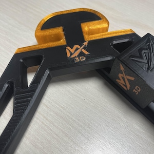 Può includere: Oggetto nero e oro stampato in 3D con la scritta "MX 3D". L'oggetto ha un design geometrico con una base rettangolare e una parte superiore curva. Le sezioni dorate hanno un aspetto traslucido.