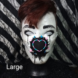 May include: A black face mask with a heart-shaped design, adorned with pink, blue, and black spikes and beads. The word "Large" is printed at the bottom of the image. The mask is on a white mannequin head with dark eye makeup.