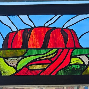 May include: Stained glass panel depicting a red rock formation against a blue sky. The artwork features vibrant colors, including red, green, yellow, and blue, and is framed in black. The design evokes a landscape scene.
