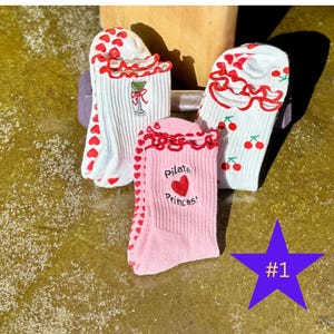 Könnte beinhalten: Drei Paar Socken mit einzigartigen Designs. Ein Paar ist rosa mit dem Text "Pilate Princess" und einem Herz. Ein weiteres Paar ist weiß mit roten Herzen, und das dritte Paar ist weiß mit Kirschen. Alle haben einen roten Rüschenbesatz.