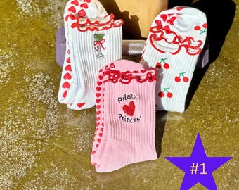 Puede incluir: Tres pares de calcetines con diseños únicos. Un par es rosa con el texto "Pilate Princess" y un corazón. Otro par es blanco con corazones rojos, y el tercer par es blanco con cerezas. Todos tienen un ribete rojo con volantes.