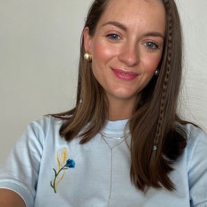 May include: A light blue t-shirt with an embroidered floral design featuring a blue flower and wheat stalks. The person is wearing gold earrings with a pearl and a silver necklace. Braids are visible in the hair.