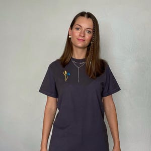 May include: Dark gray t-shirt with a small embroidered floral design in yellow, blue, and green. The shirt has short sleeves and a crew neck. The model is wearing a silver necklace and earrings.