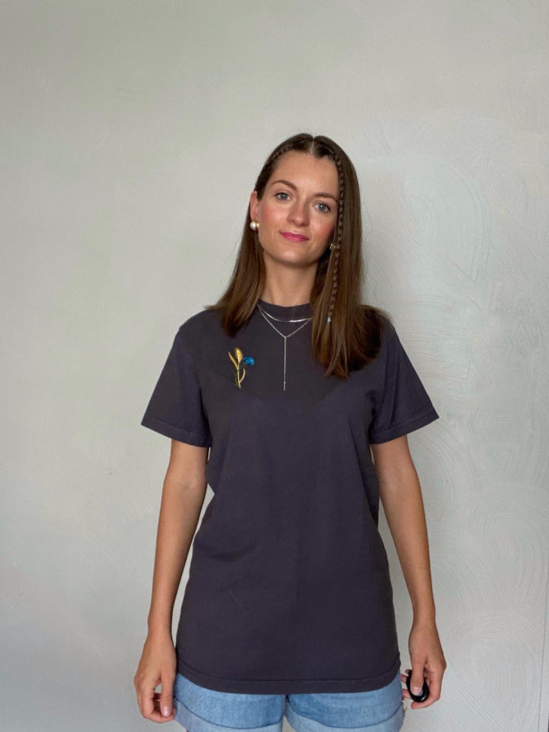 May include: Dark gray t-shirt with a small embroidered floral design in yellow, blue, and green. The shirt has short sleeves and a crew neck. The model is wearing a silver necklace and earrings.