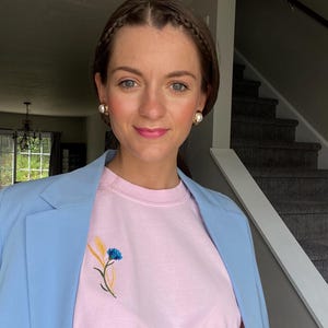 May include: A light blue blazer worn over a pink t-shirt with an embroidered floral design. The floral design features a blue flower and yellow wheat. The person is wearing gold earrings.