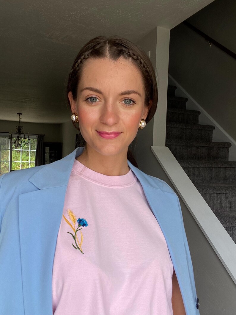 May include: A light blue blazer worn over a pink t-shirt with an embroidered floral design. The floral design features a blue flower and yellow wheat. The person is wearing gold earrings.