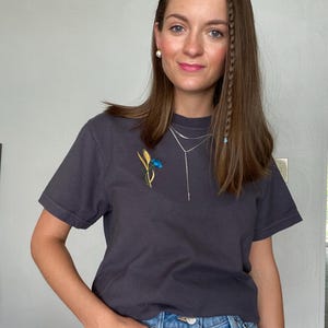 May include: Charcoal gray t-shirt with a floral embroidery of blue and yellow flowers. The shirt is paired with blue denim jeans. A silver necklace and pearl earrings complete the look. The person is standing in front of a light gray wall.