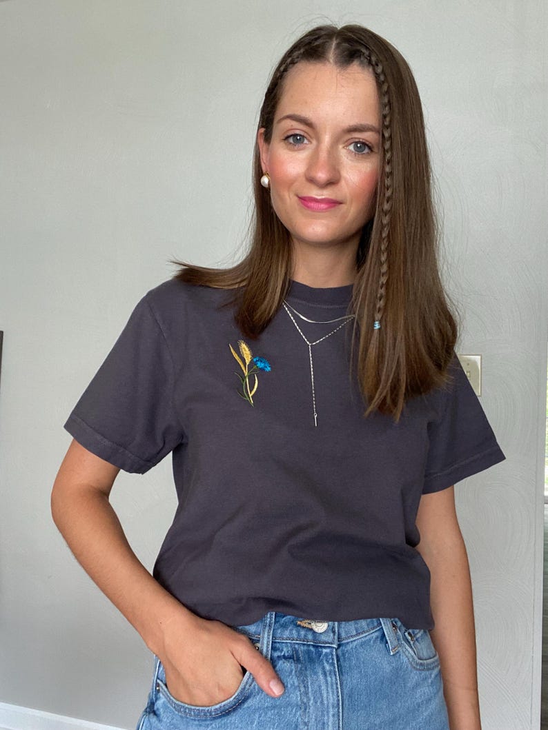 May include: Charcoal gray t-shirt with a floral embroidery of blue and yellow flowers. The shirt is paired with blue denim jeans. A silver necklace and pearl earrings complete the look. The person is standing in front of a light gray wall.