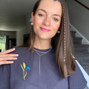 May include: A dark gray t-shirt with an embroidered blue and yellow flower design. The shirt is paired with a silver necklace. The person is wearing pearl earrings and has braided hair.