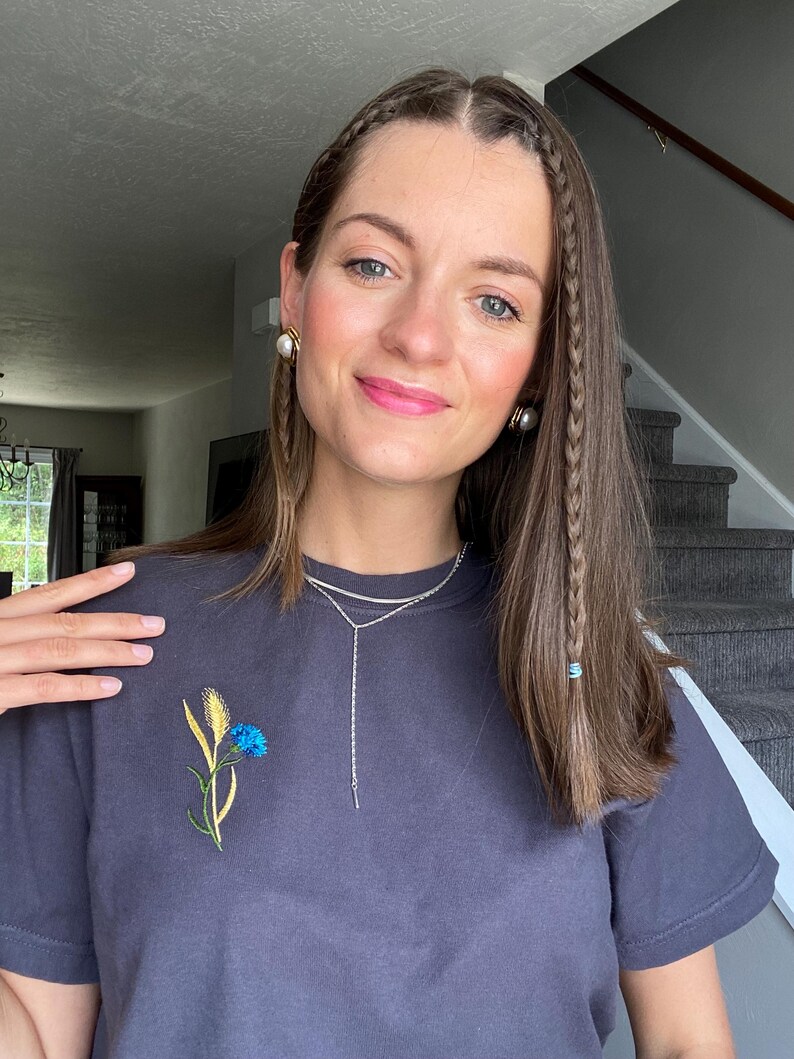 May include: A dark gray t-shirt with an embroidered blue and yellow flower design. The shirt is paired with a silver necklace. The person is wearing pearl earrings and has braided hair.