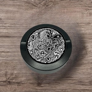 May include: A black round object with a detailed white illustration of a horse, cowboy, and floral design. The image is centered on a wooden surface, showcasing a Western theme.