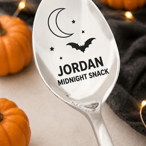 May include: A polished silver metal spoon featuring a black engraved design of a crescent moon, stars, and a flying bat. Below the graphic, the text reads JORDAN followed by the words MIDNIGHT SNACK in a clean, sans-serif capital font.
