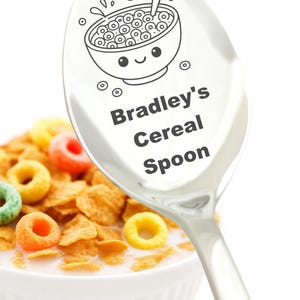 May include: A shiny silver spoon with a cartoon bowl of cereal design and the text "Bradley's Cereal Spoon." The spoon is positioned over a bowl of cereal with colorful O-shaped cereal pieces and flakes in milk.