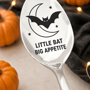 May include: A polished silver-toned metal spoon featuring a black graphic of a bat flying in front of a crescent moon with three small stars. The text "LITTLE BAT BIG APPETITE" is printed in black capital letters below the moon and bat design.