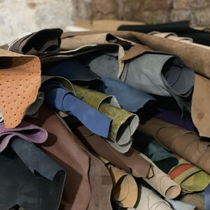 May include: A pile of various leather hides in shades of brown, tan, blue, green, and purple. Some hides are rolled, while others are flat, showcasing different textures and finishes. The leather appears to be of high quality.