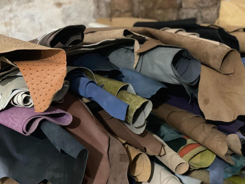 May include: A pile of various leather hides in shades of brown, tan, blue, green, and purple. Some hides are rolled, while others are flat, showcasing different textures and finishes. The leather appears to be of high quality.