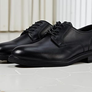May include: A pair of black leather dress shoes with laces. The shoes have a classic design with a rounded toe and a low heel. The shoes are displayed on a white tiled surface.