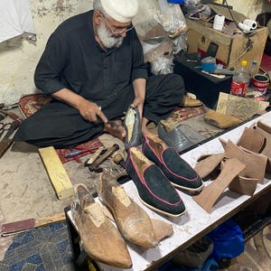 May include: A shoemaker at work, surrounded by tools and materials. Several pairs of black suede loafers with pink trim are displayed, alongside wooden shoe forms and leather pieces. The artisan is seated, focused on crafting a shoe.