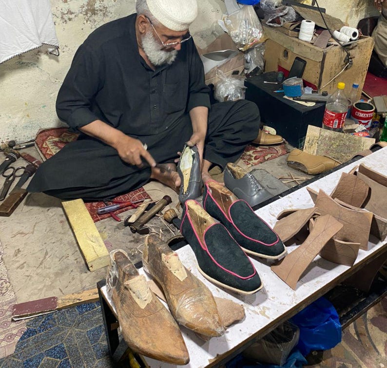 May include: A shoemaker at work, surrounded by tools and materials. Several pairs of black suede loafers with pink trim are displayed, alongside wooden shoe forms and leather pieces. The artisan is seated, focused on crafting a shoe.