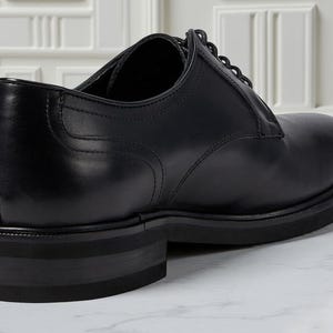 May include: A black leather dress shoe with a rounded toe and a thick sole. The shoe features a lace-up design and a low heel. The shoe is made of smooth, polished leather and is suitable for formal occasions.