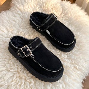 May include: A pair of black suede slip-on clogs featuring a thick black rubber sole, a plush black faux fur lining, and a decorative silver-tone metal buckle strap across the top with prominent white contrast stitching along the edges.