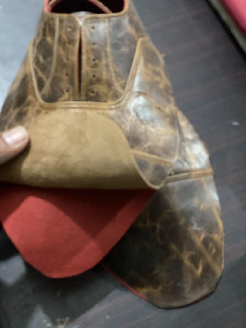 May include: Close-up of a brown leather shoe with a distressed finish. The shoe's interior is red, and the leather has a worn, textured appearance. The shoe is partially open, revealing the inner lining.