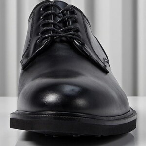 May include: A close-up shot of a black leather dress shoe with laces. The shoe has a glossy finish and a thick sole. The laces are neatly tied, and the shoe appears to be new.