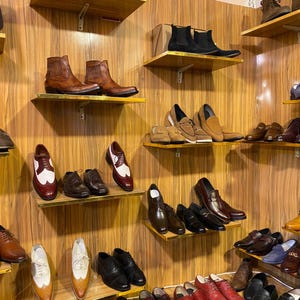 May include: A display of various men's shoes on wooden shelves. The selection includes leather boots, dress shoes, loafers, and oxfords in shades of brown, black, and tan. The shoes are arranged on multiple shelves against a wood-paneled wall.