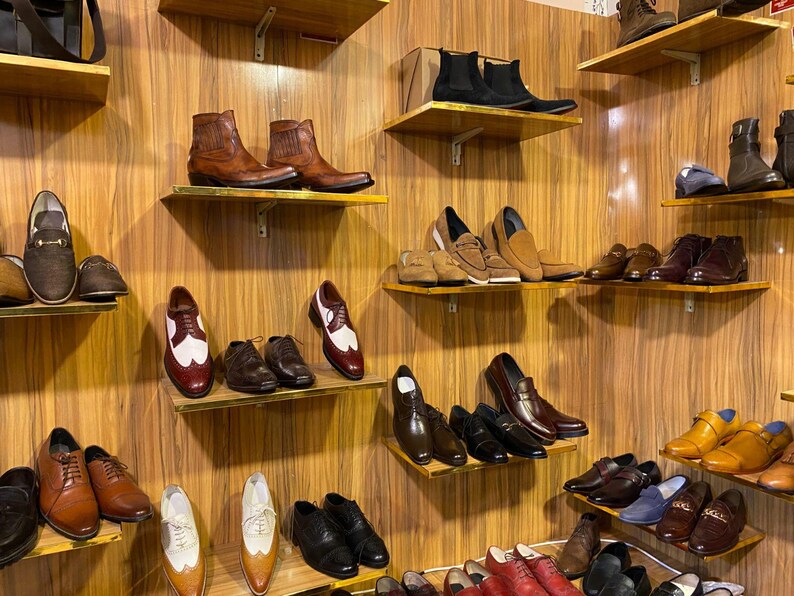 May include: A display of various men's shoes on wooden shelves. The selection includes leather boots, dress shoes, loafers, and oxfords in shades of brown, black, and tan. The shoes are arranged on multiple shelves against a wood-paneled wall.