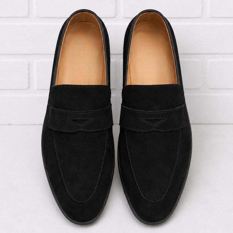 May include: A pair of black suede penny loafers. The shoes have a classic design with a strap across the vamp and a rounded toe. The interior is a light tan color, and the shoes are set against a white brick wall.