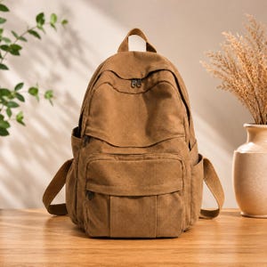 May include: A solid brown canvas backpack featuring a large main compartment with a dual zip closure, a smaller front zipped pocket, and two side pockets. The bag includes a top handle and adjustable shoulder straps for carrying.