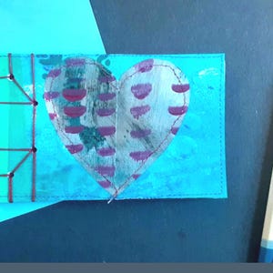 May include: A handmade greeting card with a heart design. The heart is silver with purple accents, stitched onto a turquoise background. Two pencils with blue tips are on the right. The card has a unique, artistic style.