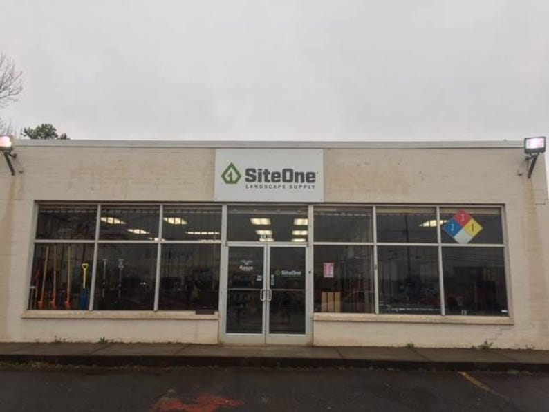 Op de afbeelding: Buitenaanzicht van een SiteOne Landscape Supply winkel. Het gebouw heeft grote glazen ramen en een dubbele deuringang. De naam en het logo van de winkel staan boven de ingang. Het gebouw is lichtbeige geschilderd.