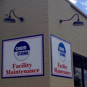 Op de afbeelding: Buitenaanzicht van een gebouwhoek met twee borden met de tekst "CHEM CLEAN Facility Maintenance". De borden hebben een witte achtergrond met blauwe en rode randen. Twee zwarte zwanenhalslampen zijn boven de borden gemonteerd.