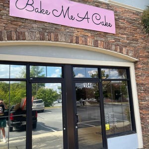 Op de afbeelding: Buitenaanzicht van een bakkerij met een roze bord met de tekst "Bake Me A Cake" in cursieve letters. De gevel heeft grote glazen deuren en ramen, met het nummer 104 boven de ingang. Het gebouw is gemaakt van baksteen en steen.