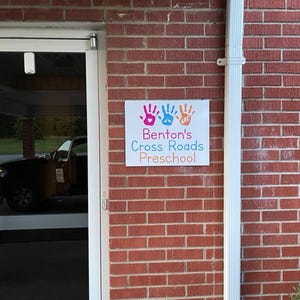 Op de afbeelding: Een bord voor Benton's Cross Roads Preschool is bevestigd aan een rode bakstenen muur. Het bord toont kleurrijke handafdrukken en de naam van de kleuterschool in een speels lettertype. Een witte regenpijp loopt verticaal langs de rechterkant van de muur.