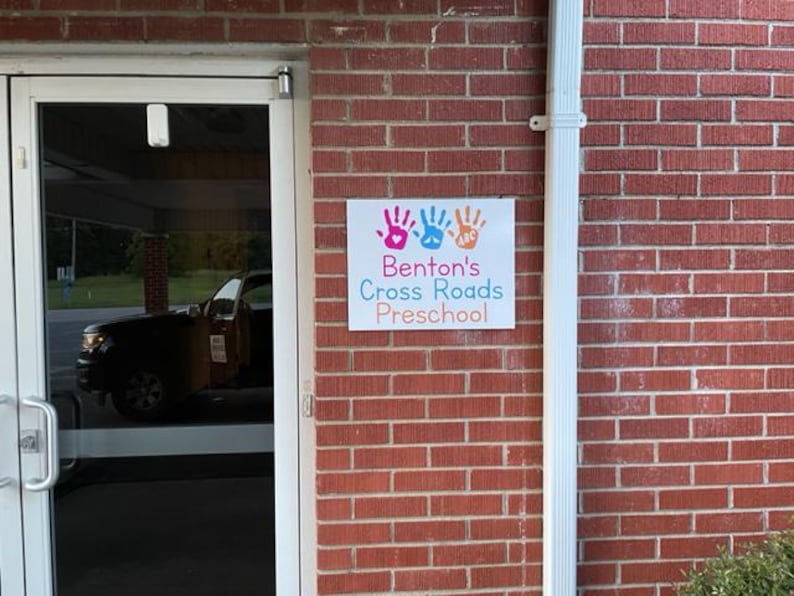 Op de afbeelding: Een bord voor Benton's Cross Roads Preschool is bevestigd aan een rode bakstenen muur. Het bord toont kleurrijke handafdrukken en de naam van de kleuterschool in een speels lettertype. Een witte regenpijp loopt verticaal langs de rechterkant van de muur.