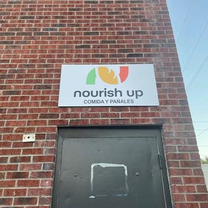 Op de afbeelding: Een rechthoekig bord met de grijze woorden "nourish up" en een kleurrijk logo erboven is op een bakstenen muur gemonteerd. Onder de tekst staat de zin "COMIDA Y PA&Ntilde;ALES" in het grijs. Een zwarte deur staat eronder.