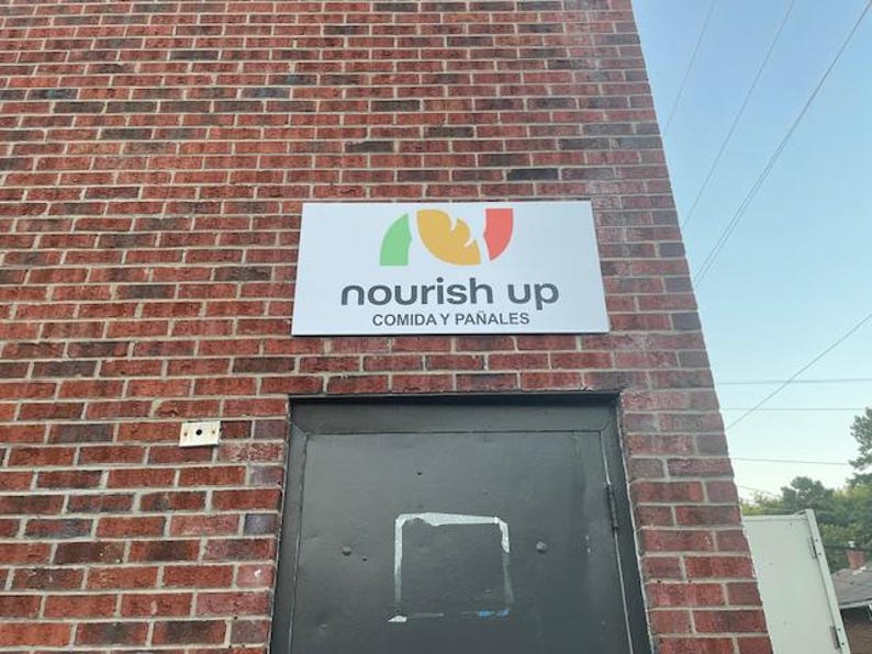 Op de afbeelding: Een rechthoekig bord met de grijze woorden "nourish up" en een kleurrijk logo erboven is op een bakstenen muur gemonteerd. Onder de tekst staat de zin "COMIDA Y PA&Ntilde;ALES" in het grijs. Een zwarte deur staat eronder.