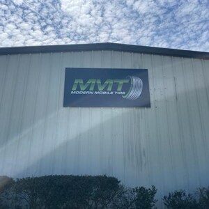 Op de afbeelding: Buitenaanzicht van een gebouw met een bord "MMT MODERN MOBILE TIRE". Het bord heeft groene tekst en een bandenafbeelding. Het gebouw heeft een buitenkant van gegolfd metaal tegen een bewolkte lucht.