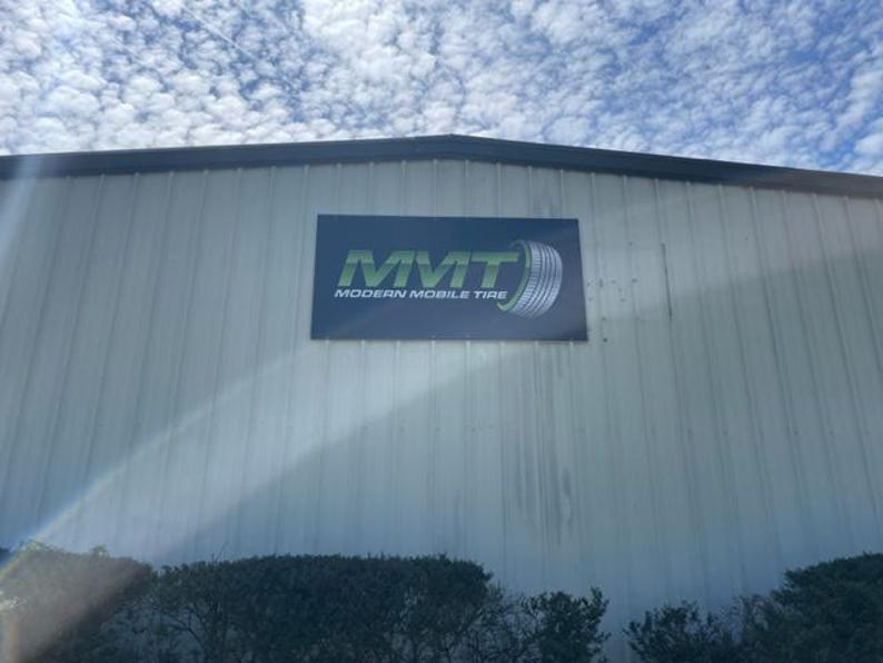 Op de afbeelding: Buitenaanzicht van een gebouw met een bord "MMT MODERN MOBILE TIRE". Het bord heeft groene tekst en een bandenafbeelding. Het gebouw heeft een buitenkant van gegolfd metaal tegen een bewolkte lucht.